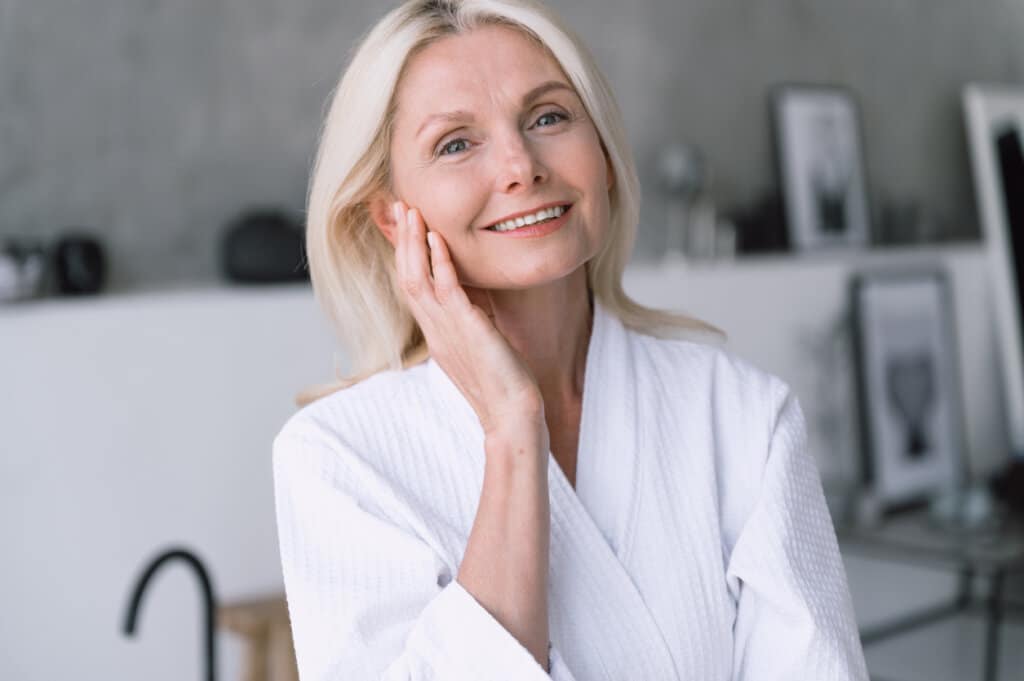 Miami’s Top Facelift Surgeon: Smiling mature woman in white robe touching cheek
